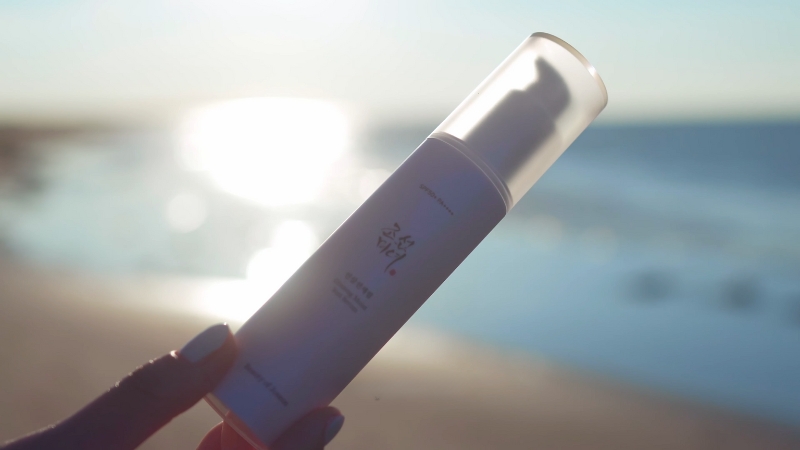 A hand holds a cruelty-free Korean skincare product with the ocean in the background