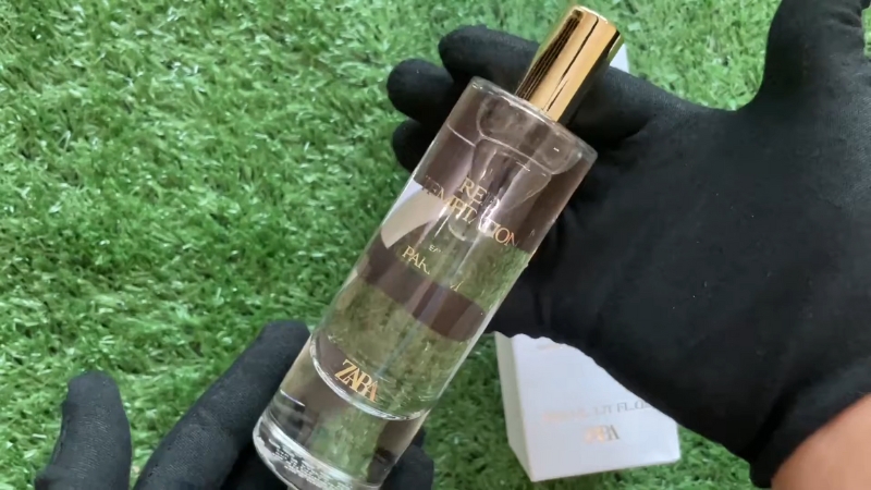 A person holding a bottle of Zara Red Temptation Eau de Parfum with a gold cap against a green background