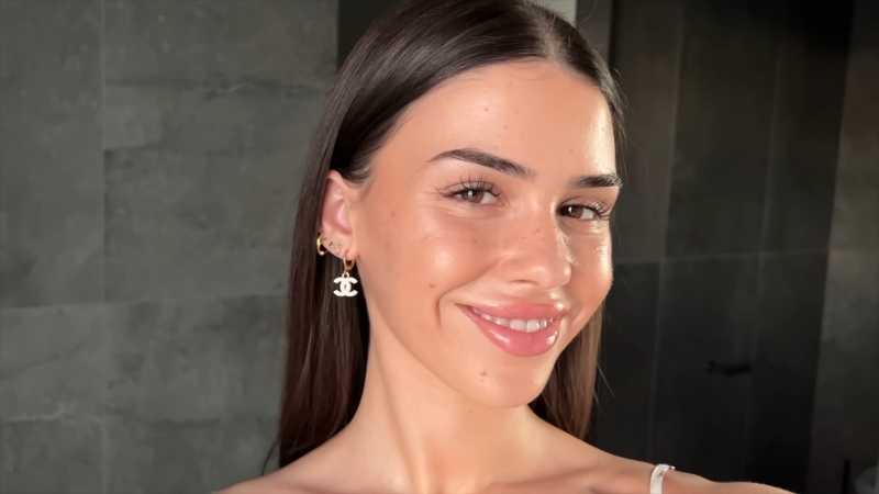 A woman with natural makeup, glossy lips, and minimal jewelry smiling