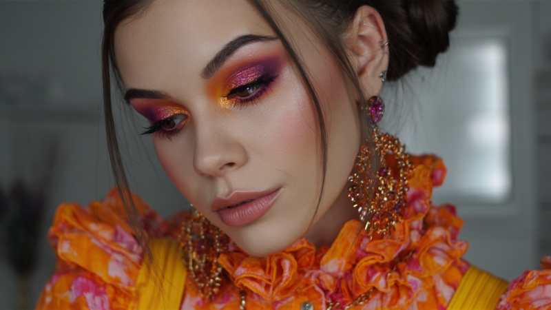 A woman wearing bold pink and orange eye makeup paired with a vibrant ruffled outfit and statement earrings
