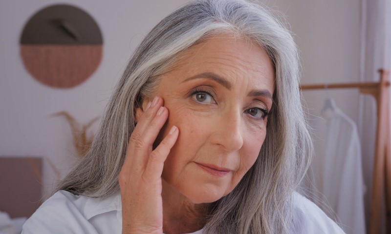 Close-up portrait of an elegant older woman with long silver hair gently touching her cheek