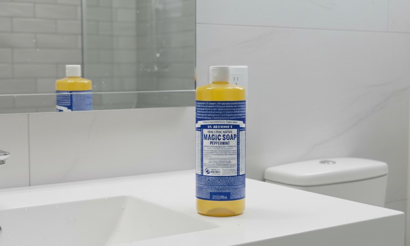 Bottle of Dr. Bronner's Magic Soap sits on a white bathroom counter beside a sink. Its reflection is visible in a mirror