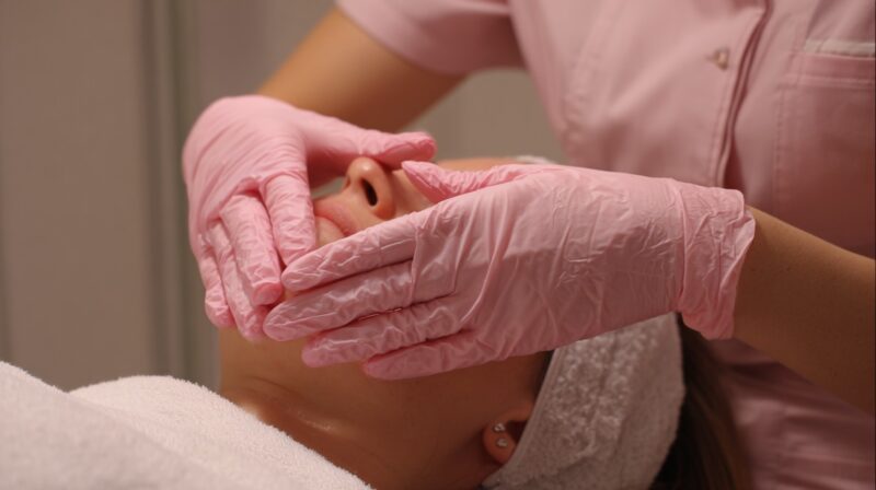 Close-up of a practitioner wearing pink gloves performing a buccal facial massage on a client lying down