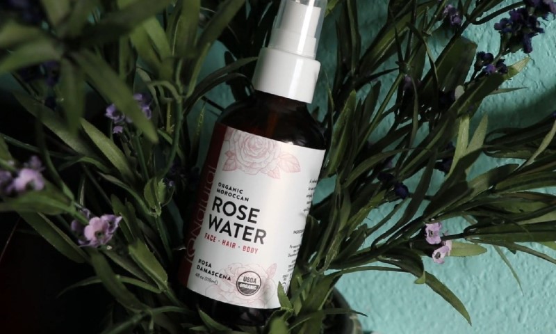A brown bottle labeled "Organic Moroccan Rose Water" sits in lush green foliage with purple flowers
