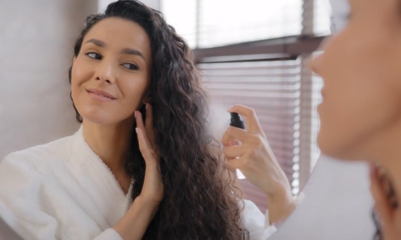 A woman with long, curly hair smiles while applying hair spray