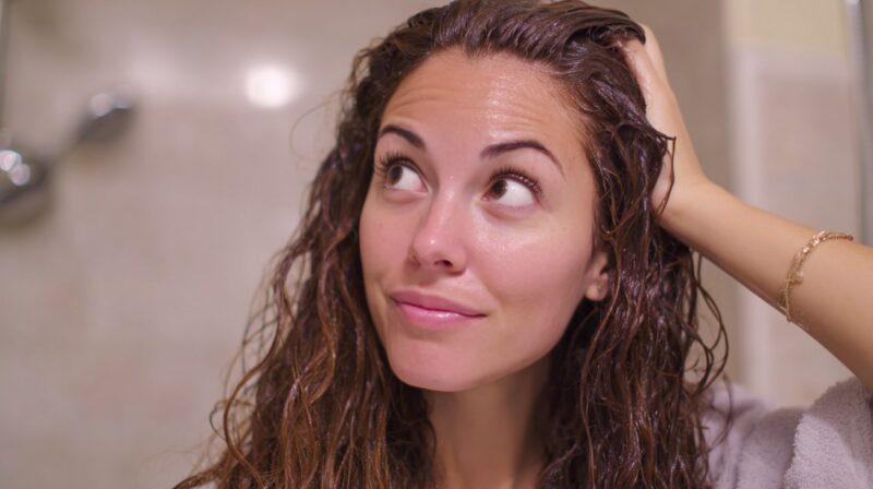 Woman with damp hair in a bathroom touching her scalp and looking upward thoughtfully