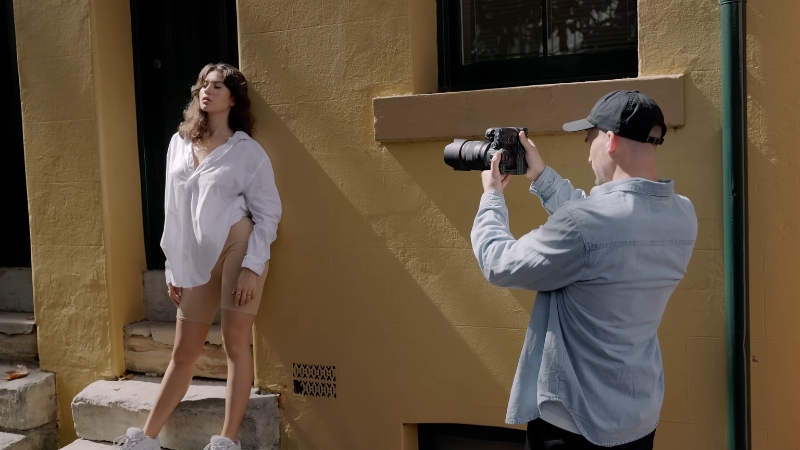 Photographer capturing a woman posing against a yellow wall while preparing for the portrait