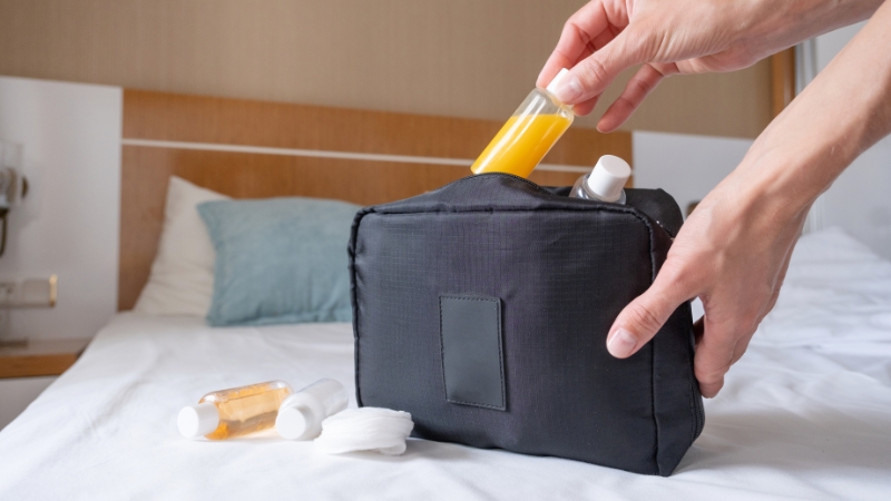 Hand placing mini skincare bottles into a travel beauty bag on a bed