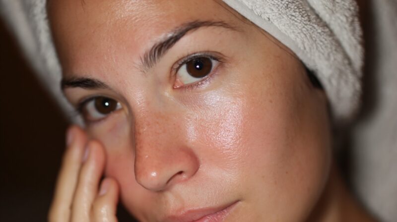 Woman with a towel wrapped around her head gently touching her cheek after skincare application