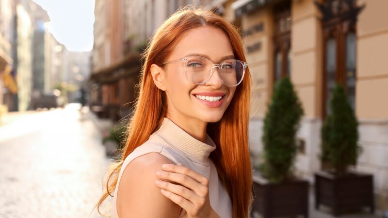 A smiling woman wears clear-frame glasses on a sunny city street
