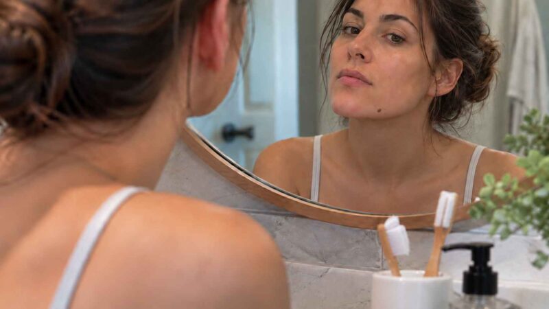 Woman examining a mole on her chin in a bathroom mirror.